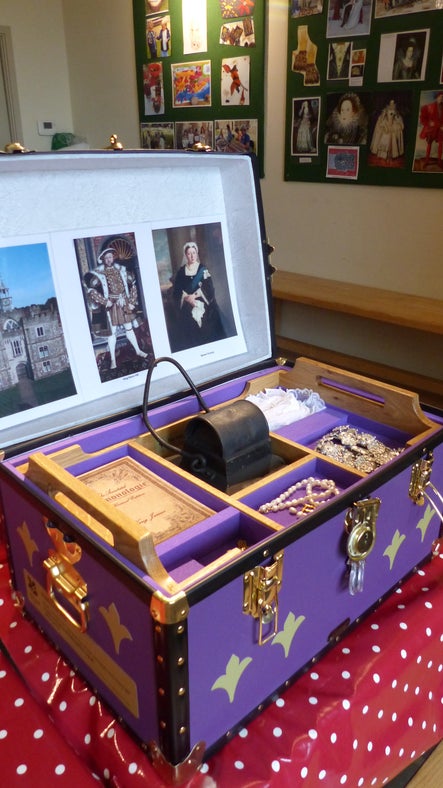 Large purple case on a table that has a red and white tablecloth on. The lid of the case is open and there are photos and items inside, including jewellery. There are photos on the wall in the background.